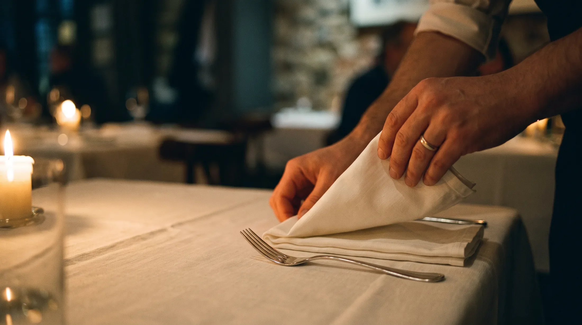 A server's hands setting a folded napkin and silverware beside a candle