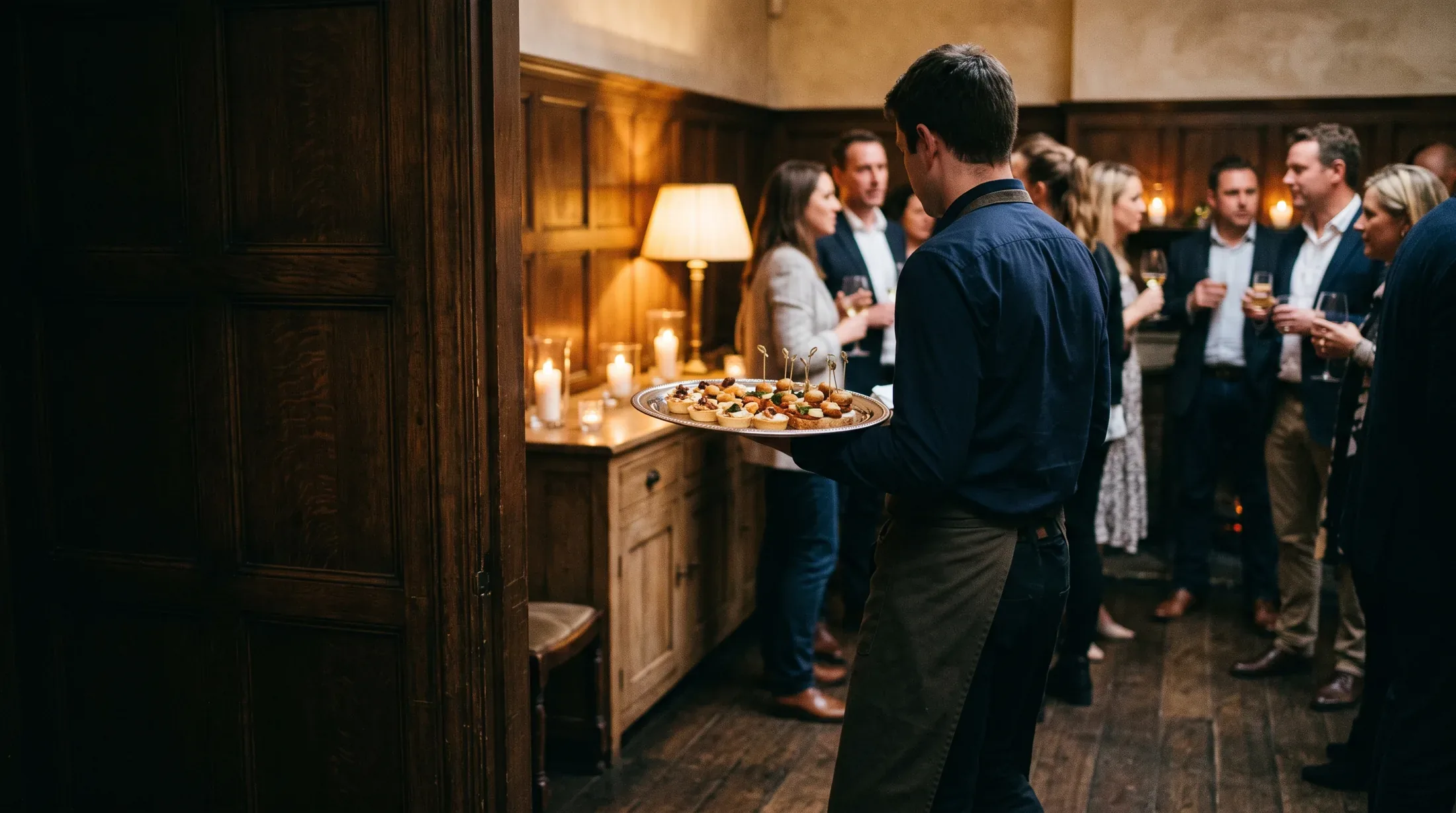 A catering server carries a tray of canapes past guests at an evening drinks reception in a warm panelled room
