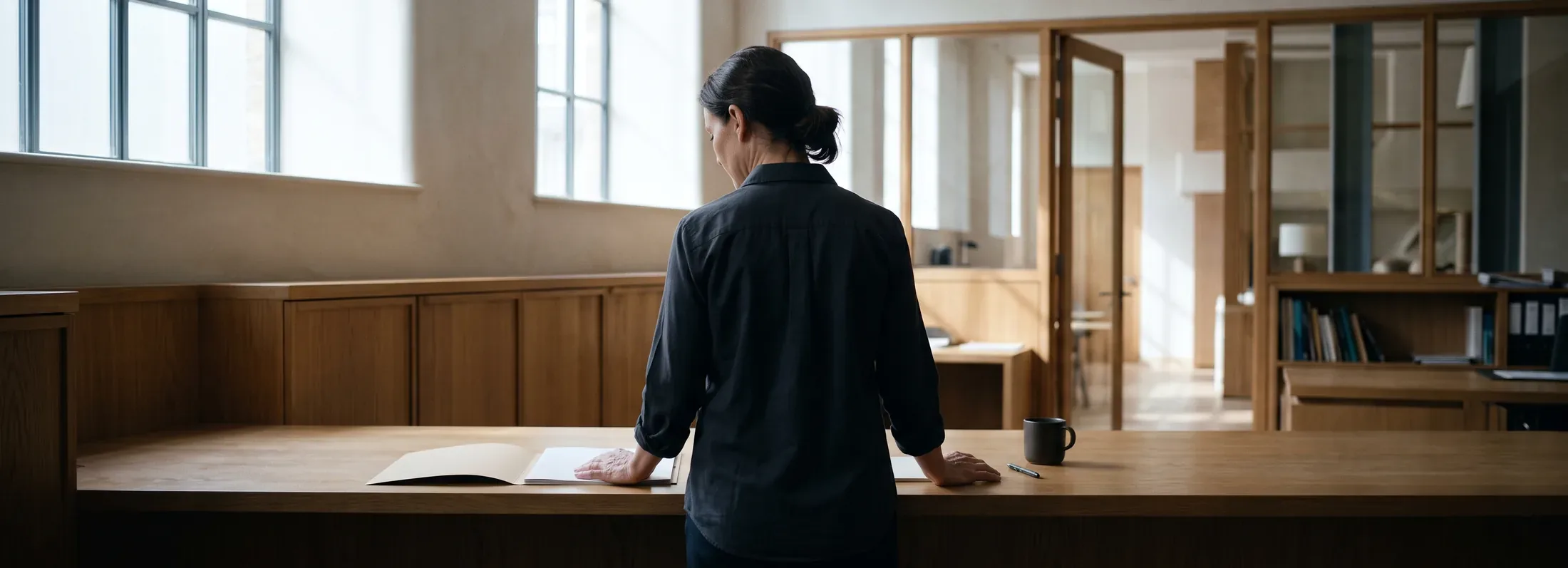 A staffing coordinator stands at a long oak counter in a daylit hotel back office, seen from behind, one hand resting on an open folder.