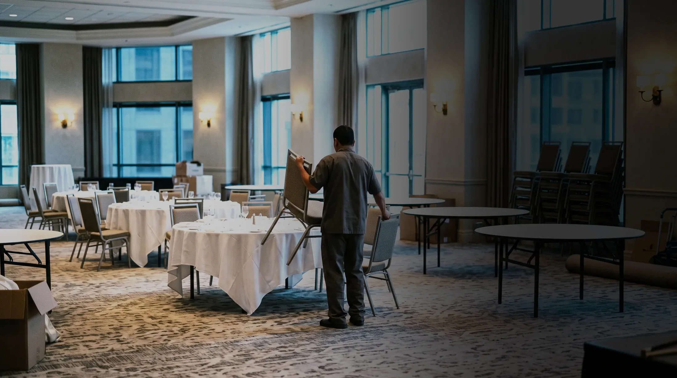 A ballroom half set for an event, one worker placing chairs while tables stand bare