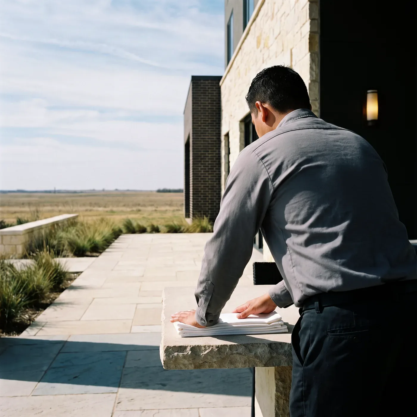 A worker straightening folded linen on a limestone table outside a Dallas property, with prairie grass and an open horizon beyond the terrace.