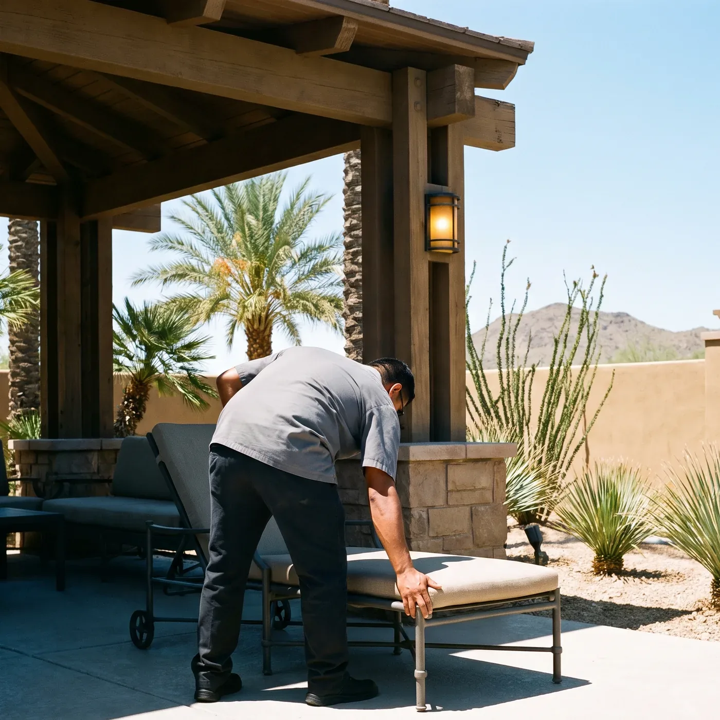 A hotel worker squaring a lounge chair under a timber ramada at a Phoenix property, with date palms, ocotillo and a desert ridge behind him.
