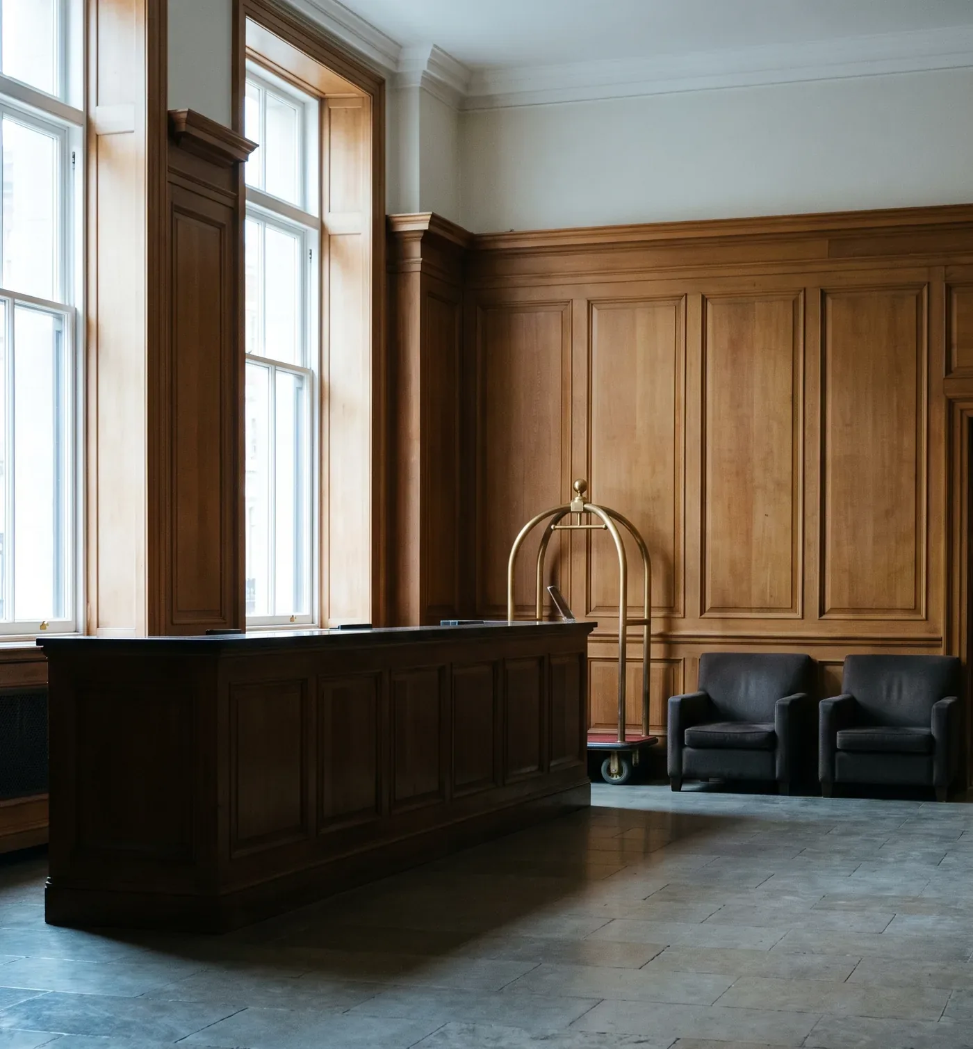 The empty reception hall of an upscale property in morning light, a timber counter and a brass luggage trolley standing ready on a worn stone floor