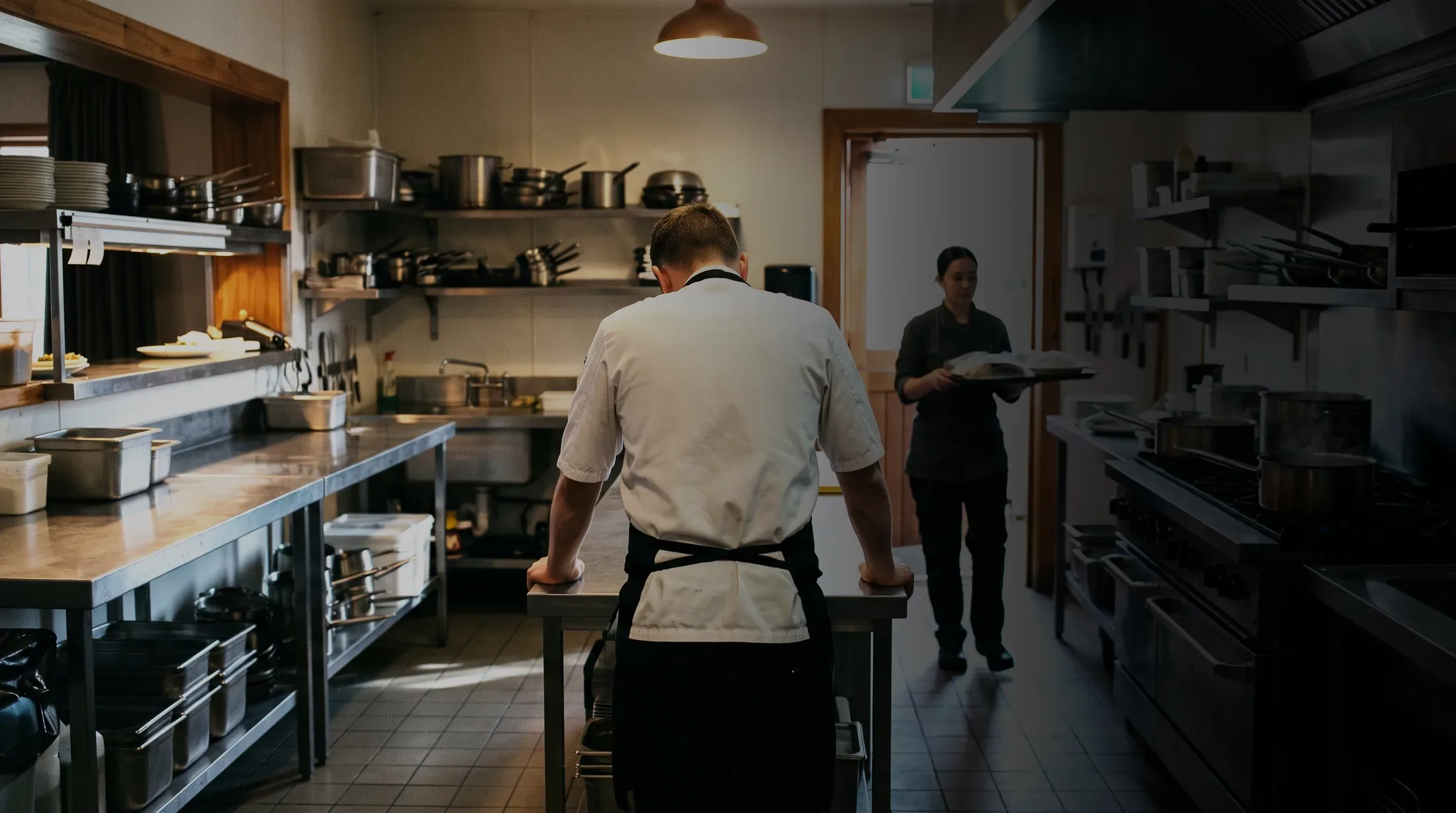 A cook braced on the pass late in a shift as a colleague carries plates out
