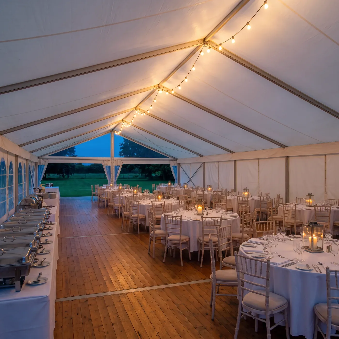 A white event marquee dressed for a gala at dusk, tables set and empty, a covered buffet line down one side and festoon lights along the ridge.