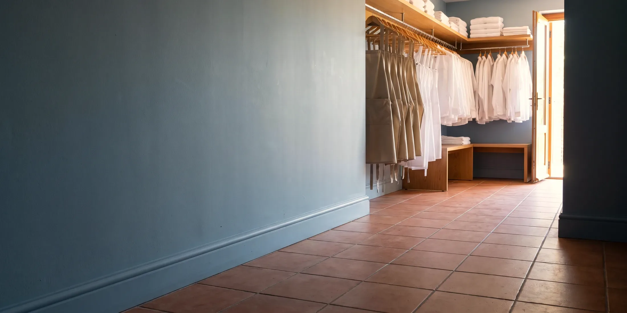 A rail of pressed uniforms on wooden hangers beside folded linen and a bench in a daylit staff uniform room at the start of the day