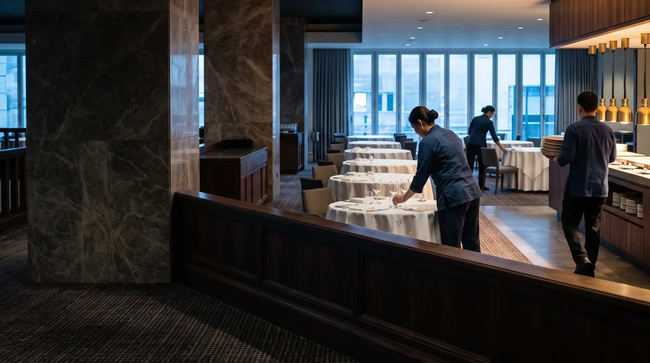 A hotel dining room being set for the morning, seen across a dark timber balustrade: one worker placing wine glasses on a linen laid table, a second straightening a chair beyond her, and a third carrying a stack of clean plates to a buffet counter under warm brass lamps