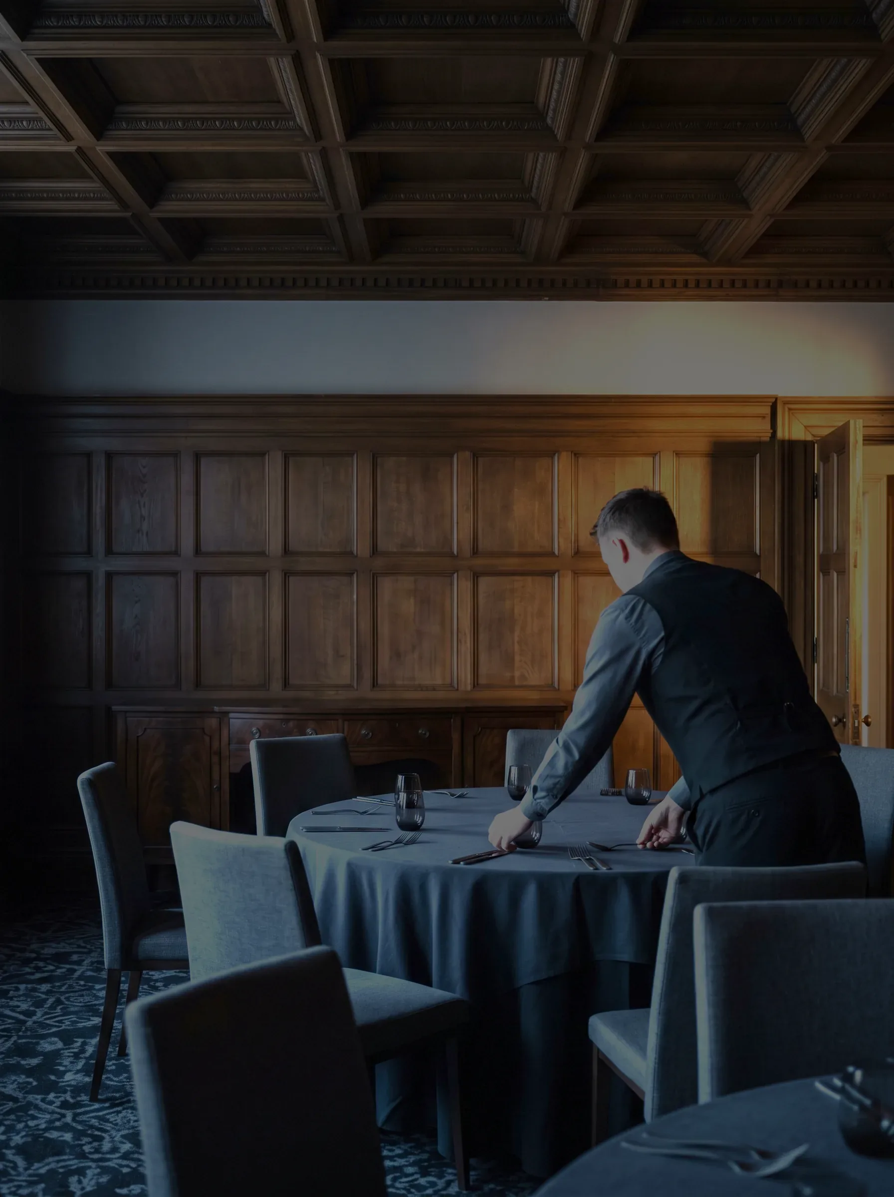 A hotel dining room being finished for service, round tables dressed with the chairs pulled in, one person in a black waistcoat seen from behind and working at a table, and the nearer chairs and the patterned carpet in shadow in the foreground.