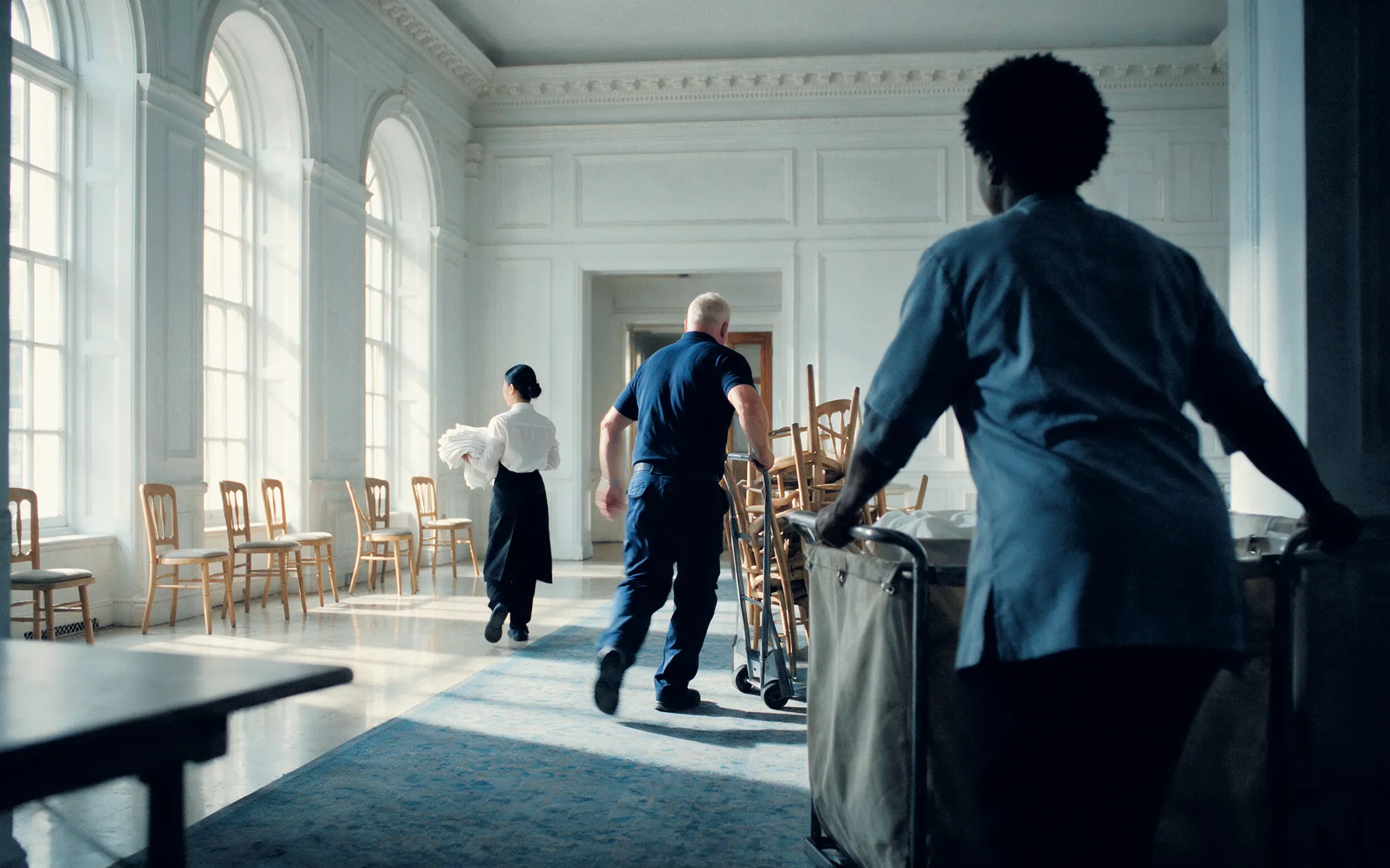 Three hotel staff turning over a pre-function room lit by tall arched windows down its left wall, one carrying a stack of folded white table linen, one mid stride behind a dolly of stacked timber banquet chairs, and one in the foreground pushing a canvas linen trolley away from the camera