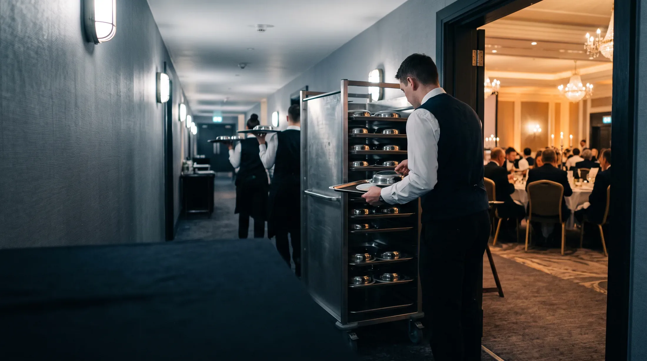 A server loading covered plates onto a tray in the service corridor beside a banquet already seated.