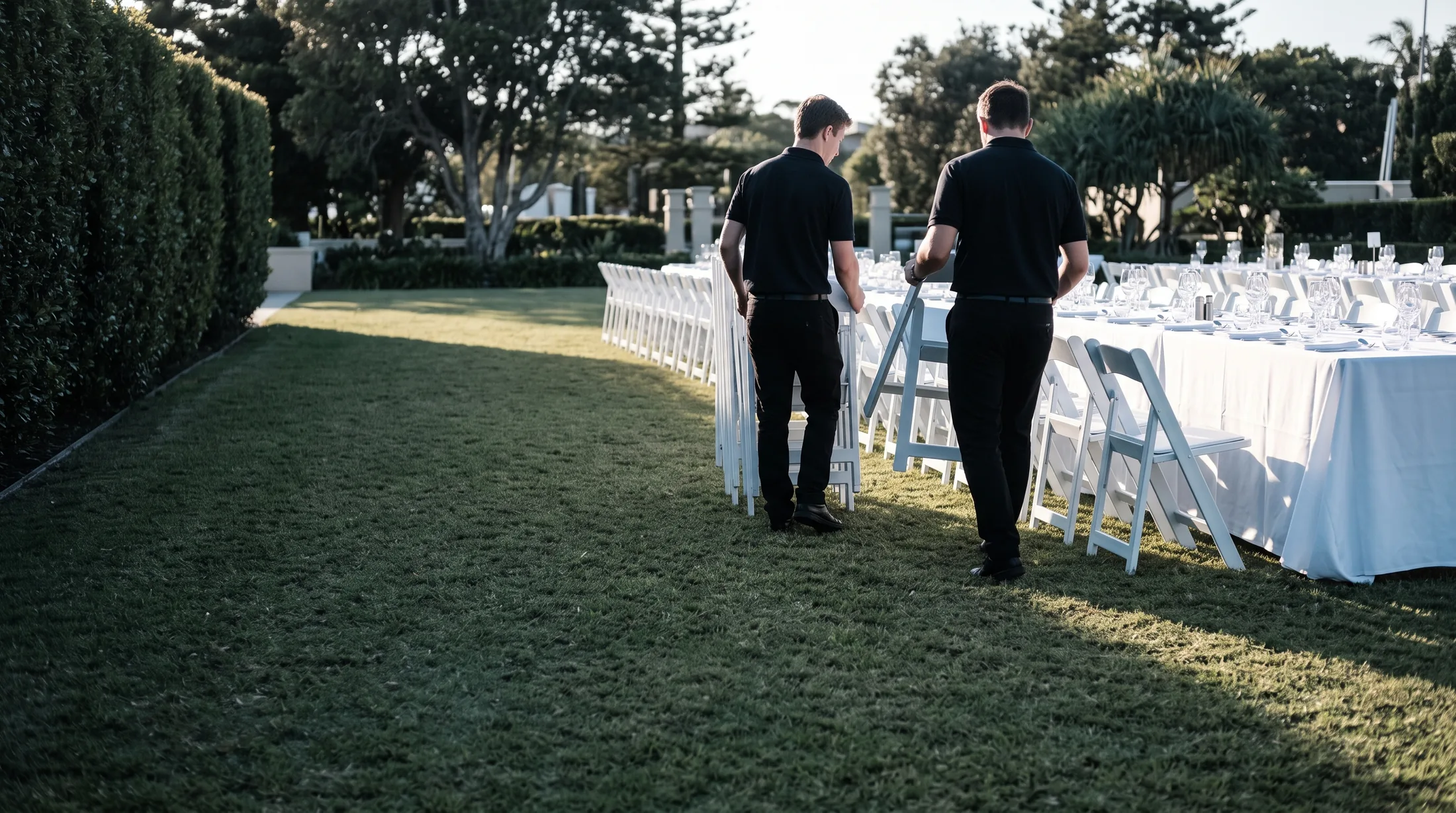 Two event staff setting white folding chairs into rows on a resort lawn, a dressed head table behind them.
