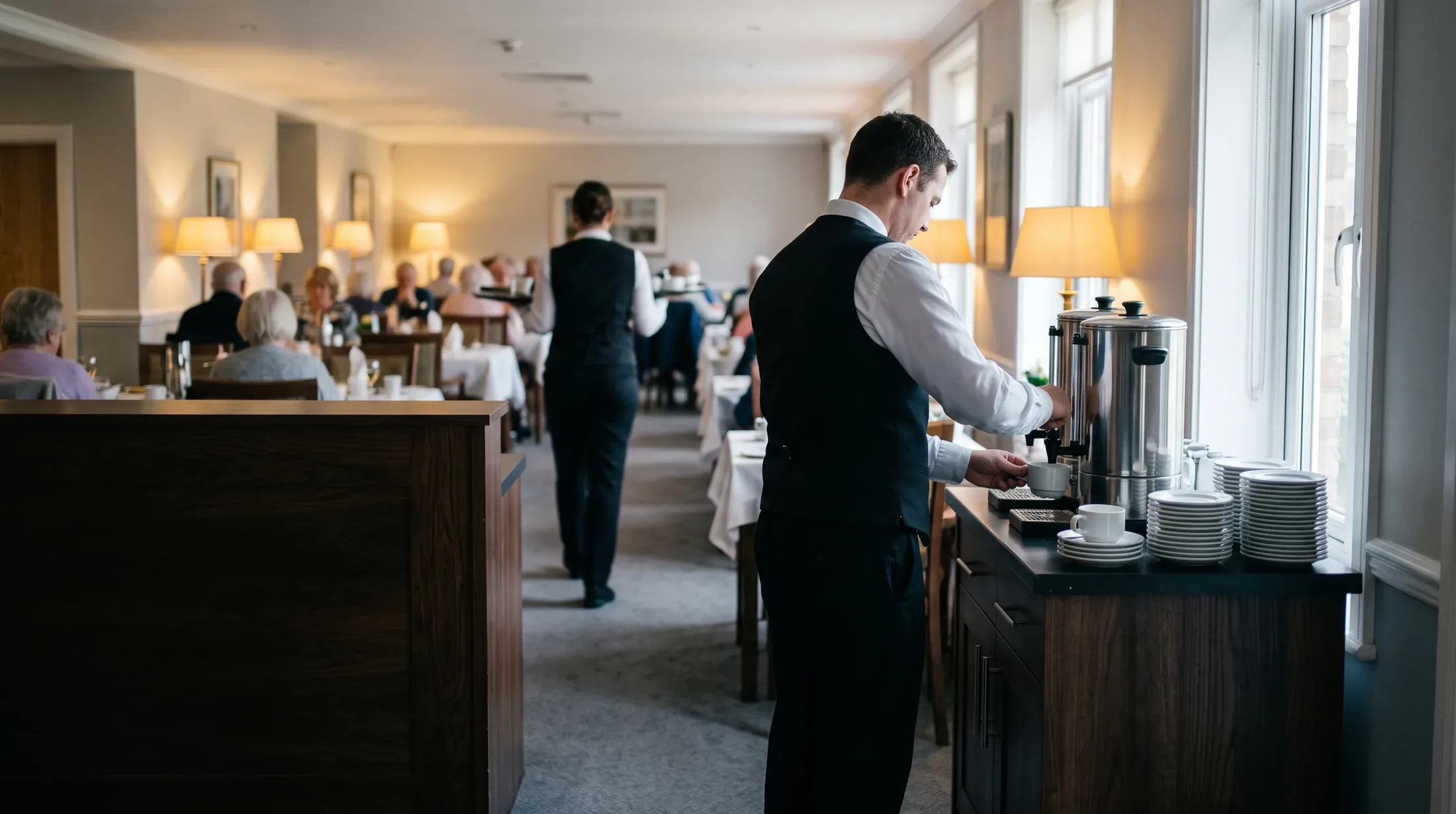 A server filling cups at a beverage station during a full midday sitting in a senior living dining room.