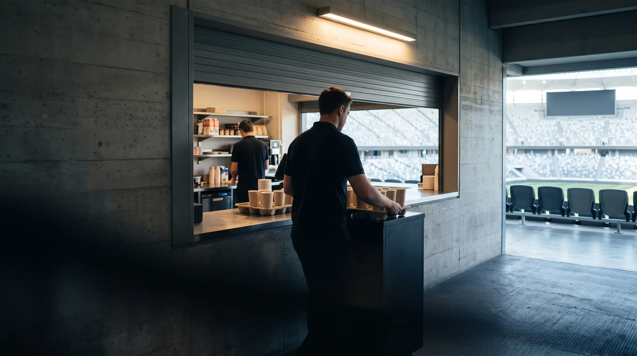 Two crew restocking a concession stand on a concourse, the empty seating bowl visible through the opening.