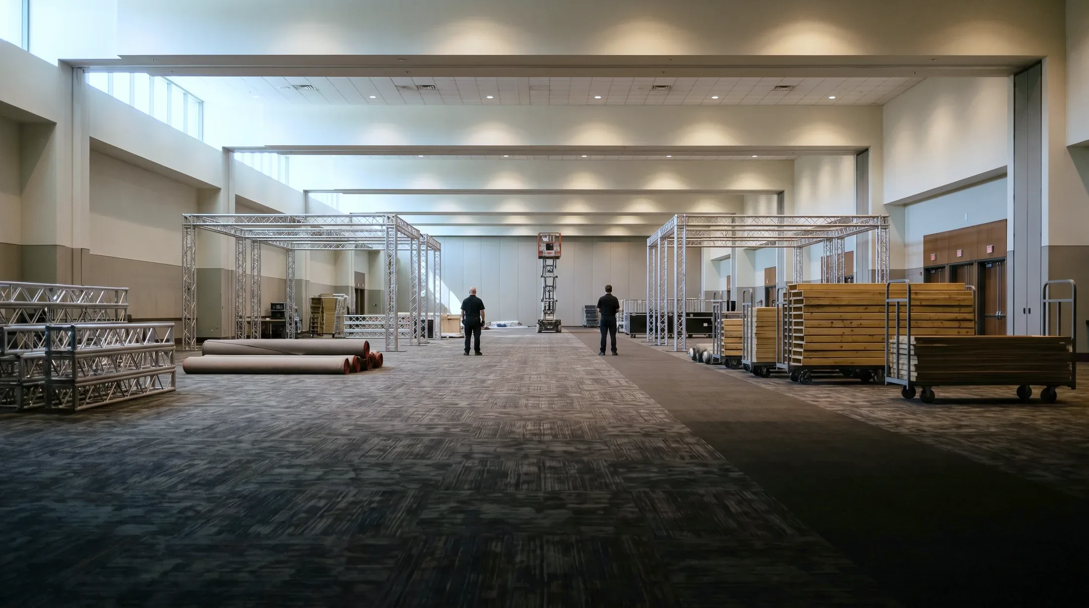 An exhibit hall between two shows, booth truss part built over rolled aisle carpet and staging carts, two workers small and far down the aisle