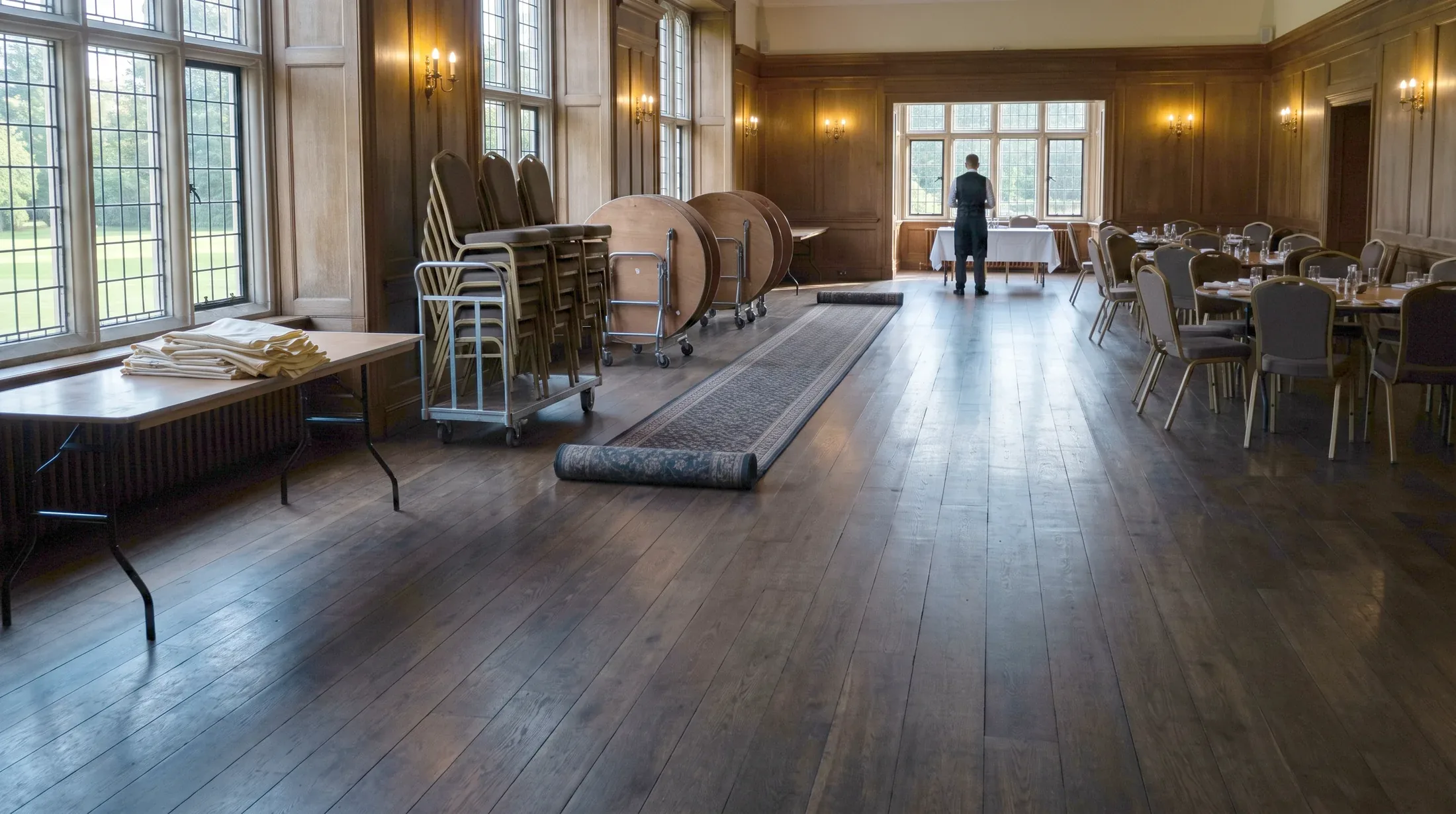 A panelled club room half struck between two events, round tables tipped on trolleys and chairs stacked beside a rolled runner, one server at the far window