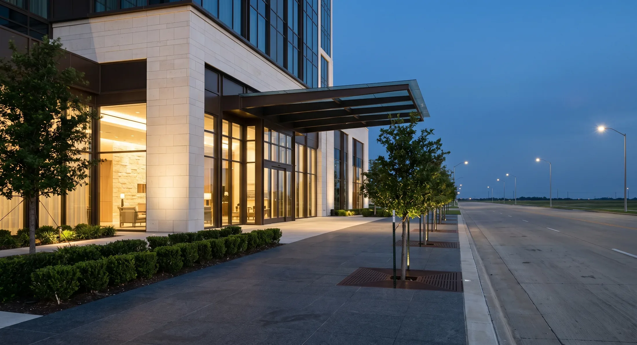 A pale limestone hotel podium on a wide empty boulevard at blue hour, warm lobby light spilling across the stone and a low flat horizon beyond