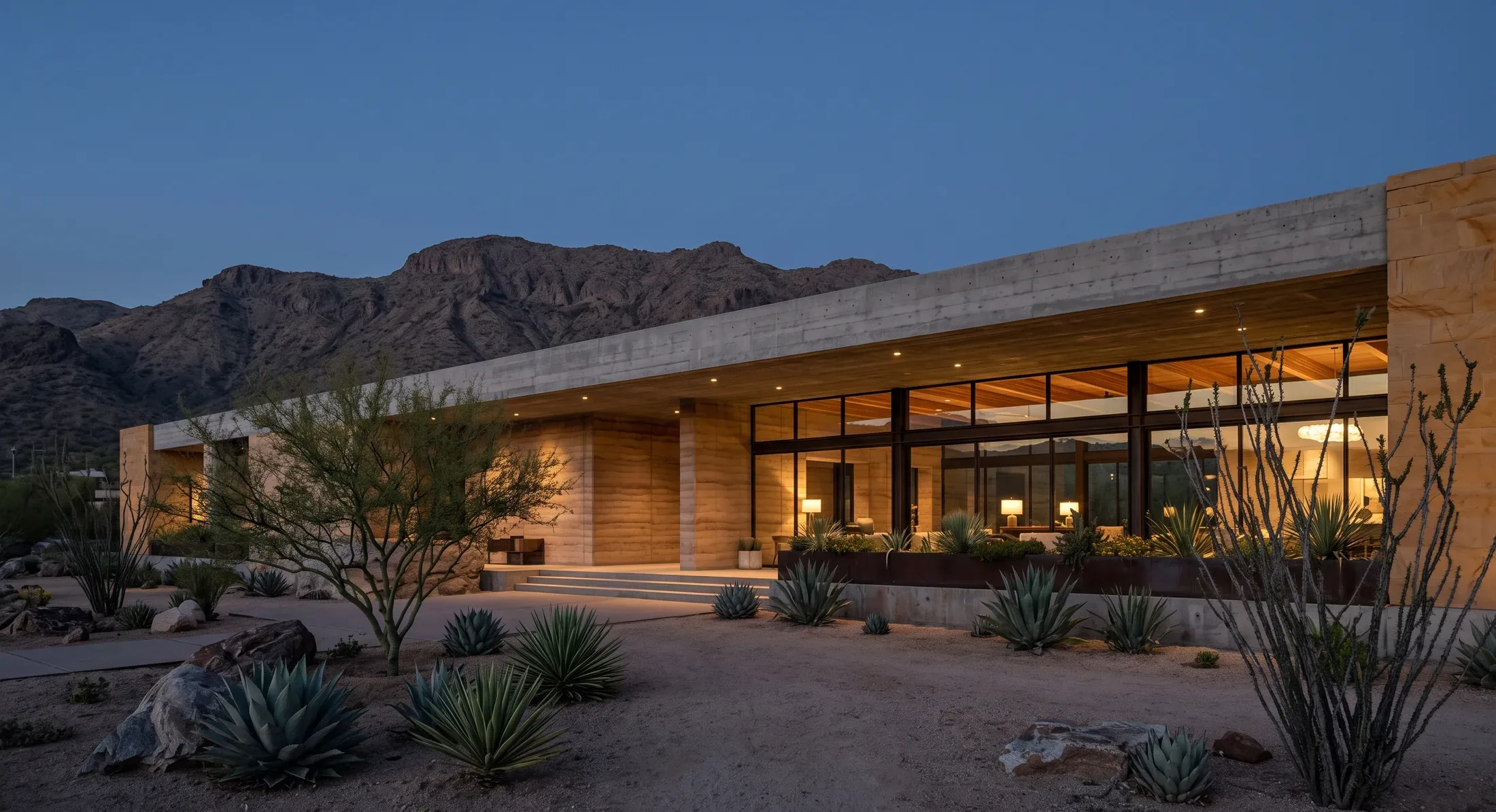 A long low desert hotel wing in rammed earth and warm sandstone at blue hour, a bare rocky ridge behind it and agave and ocotillo in the gravel foreground