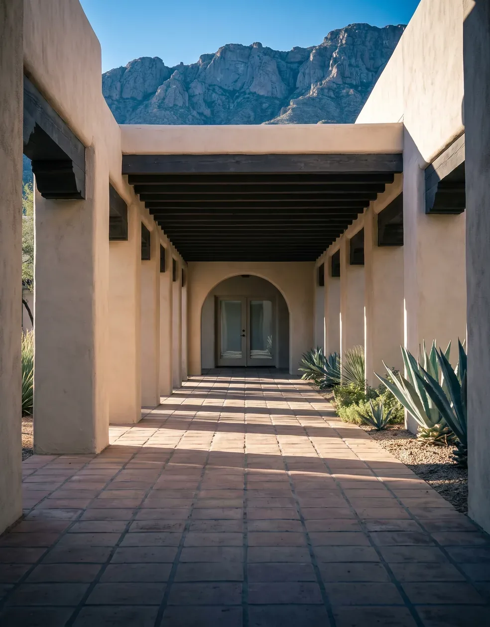 A shaded adobe arcade at a desert hotel, dark timber beams running the length of a sand stucco colonnade to an arched glass door, agave beds at the right and a granite mountain over the roofline