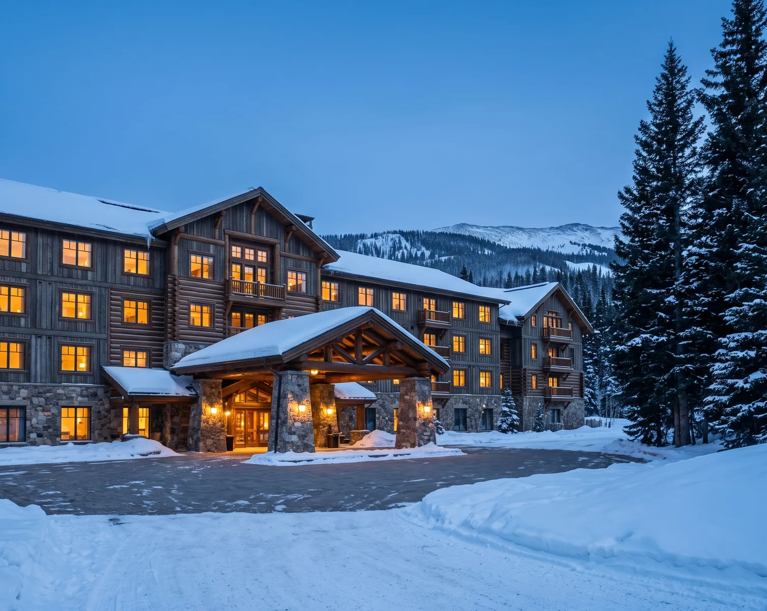 A timber and stone lodge at blue hour with snow on its roofs and banked along a cleared drive, warm light in every window and a lit porte cochere at its centre