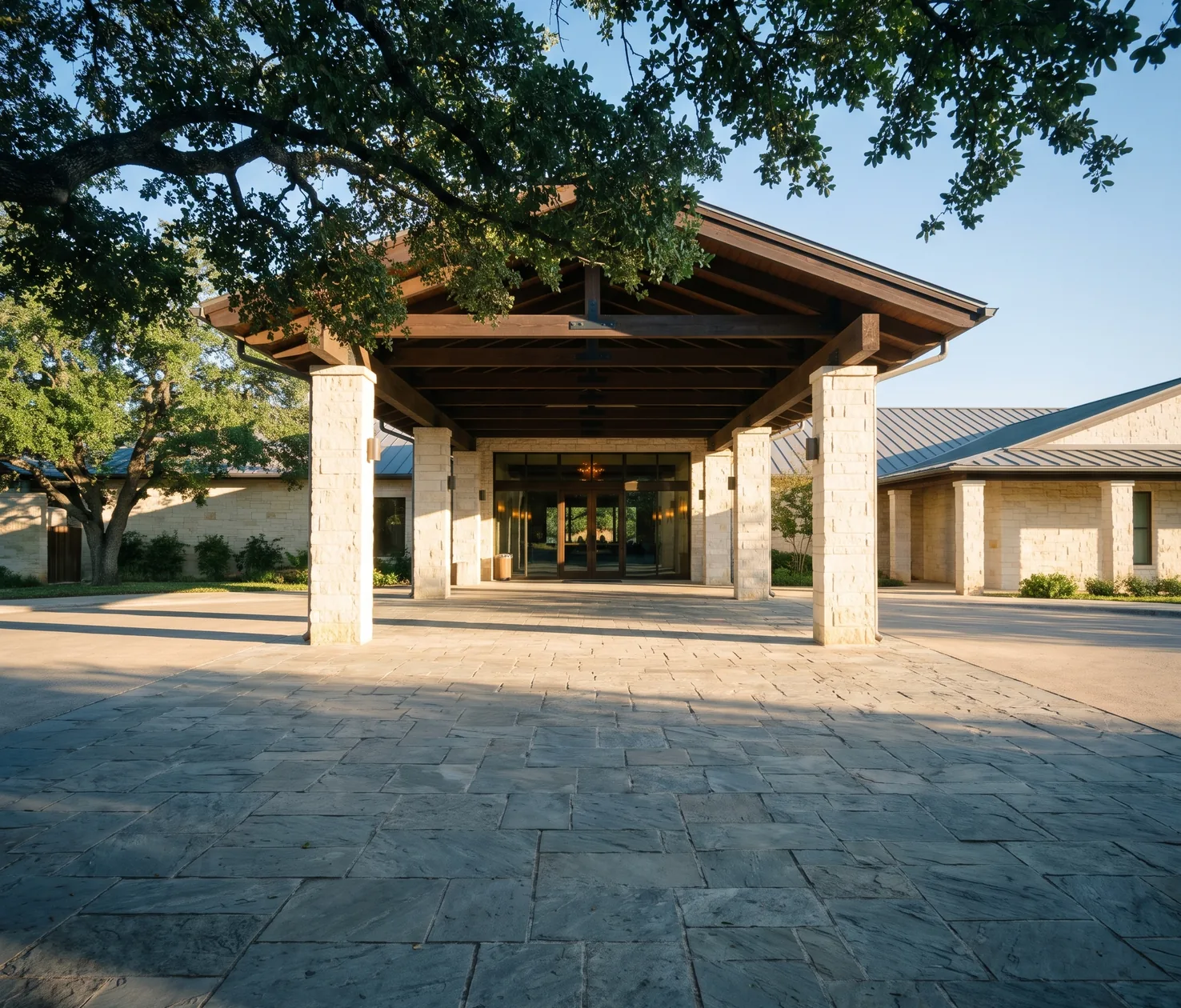 A hill country hotel arrival court, a timber gable on limestone columns over lit lobby glass, a live oak canopy across the top of the frame and a flagstone forecourt in front