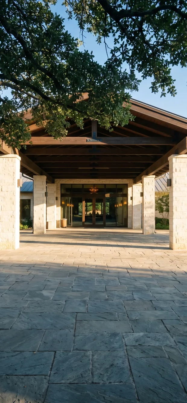 A hill country hotel arrival court, a timber gable on limestone columns over lit lobby glass, a live oak canopy across the top and a flagstone forecourt in front
