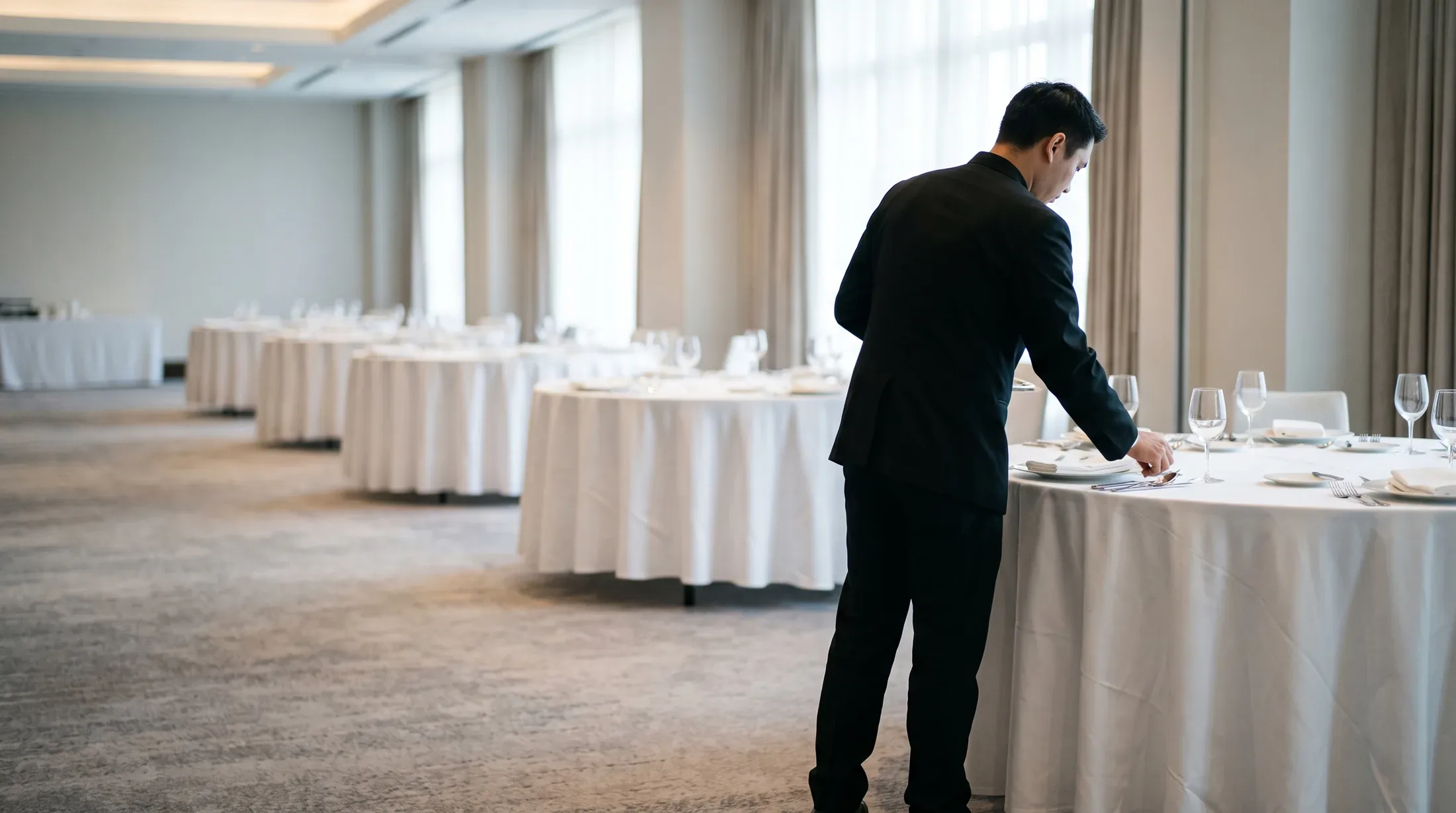 A banquet server laying cutlery at a round table dressed in white linen in a ballroom set for a function