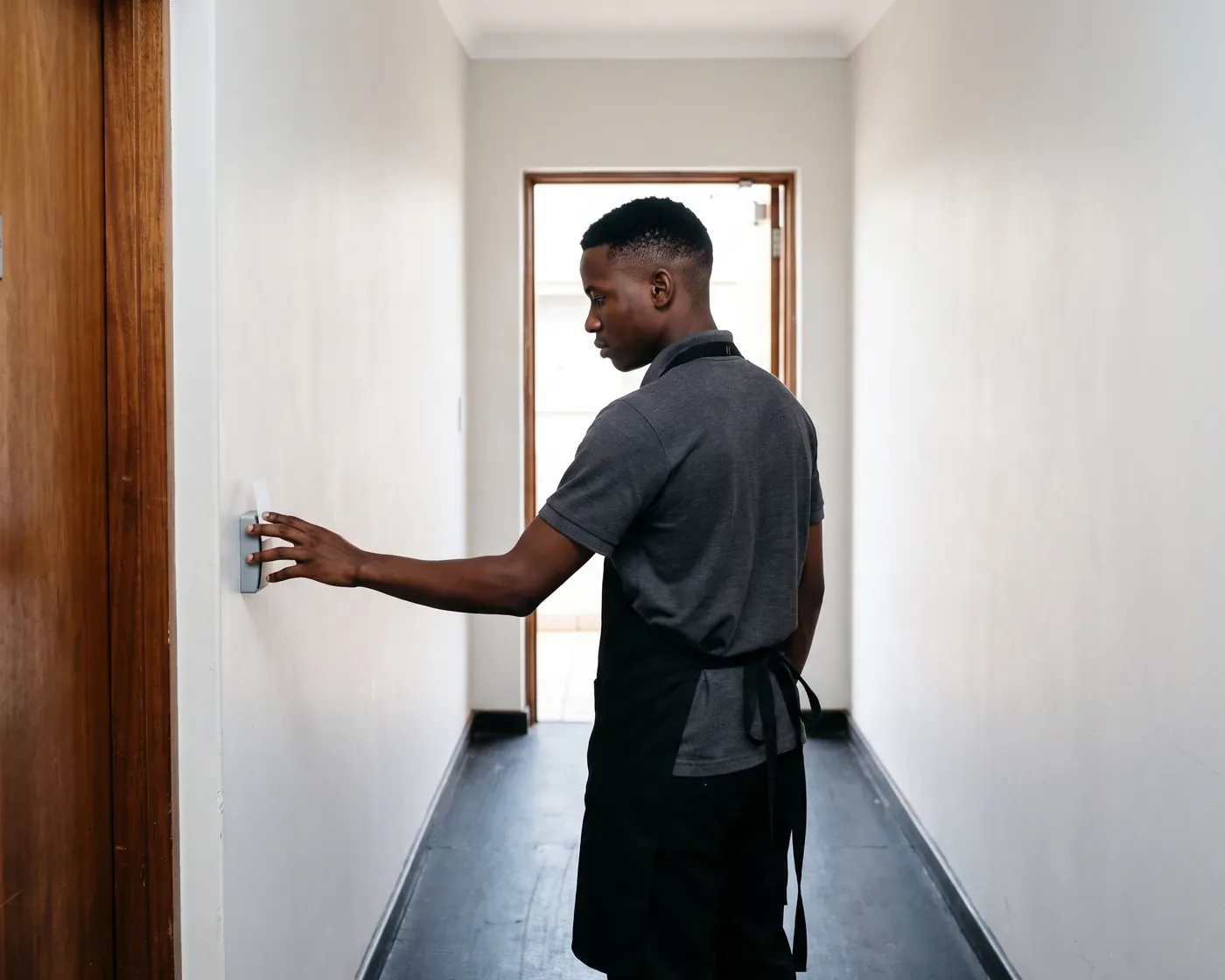 A young man in a grey polo and black apron holding a plain white key card against a small wall reader beside a timber door in a back of house corridor, daylight through the open doorway beyond him